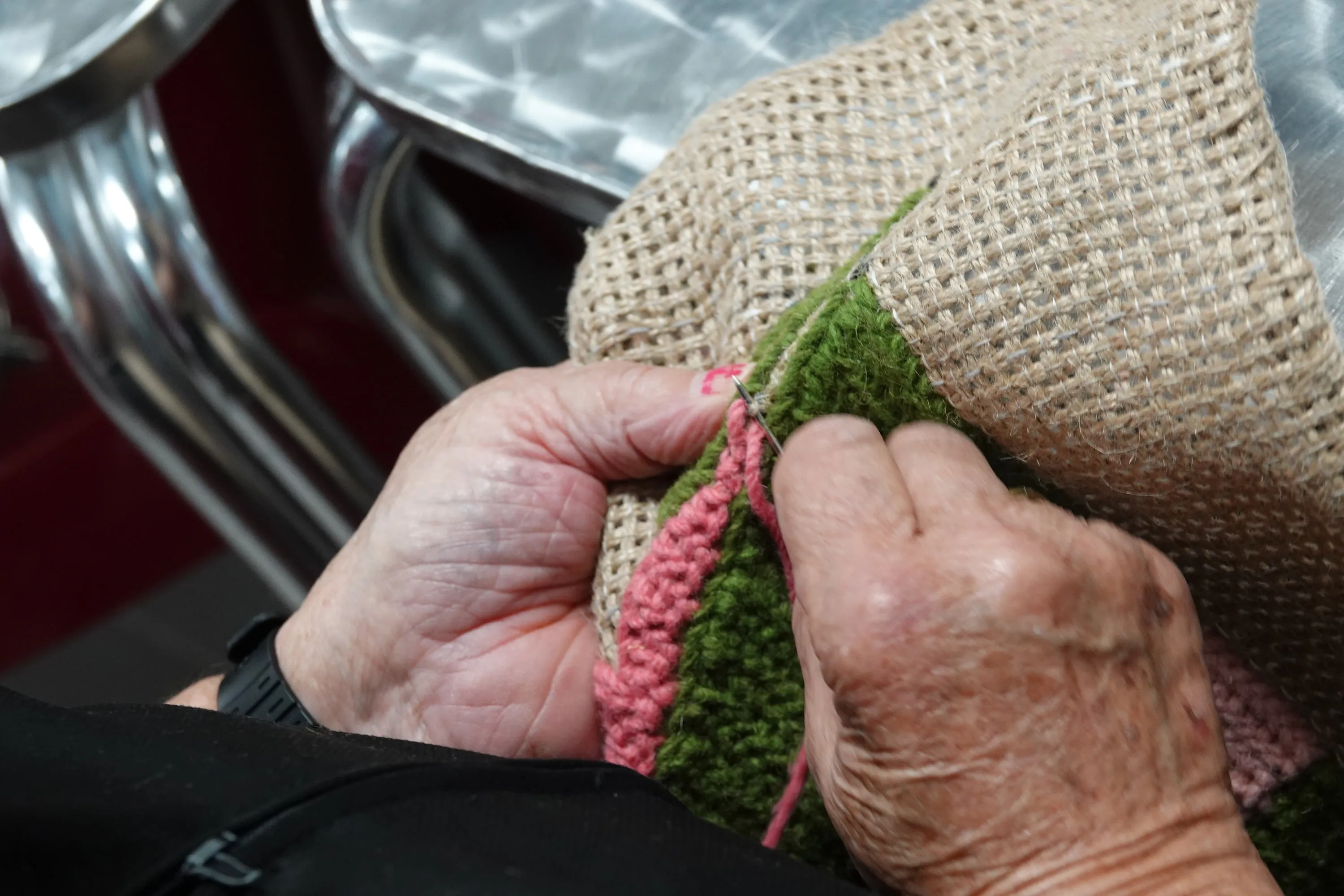 Close view of hands embroidering green and pink wool through burlap.